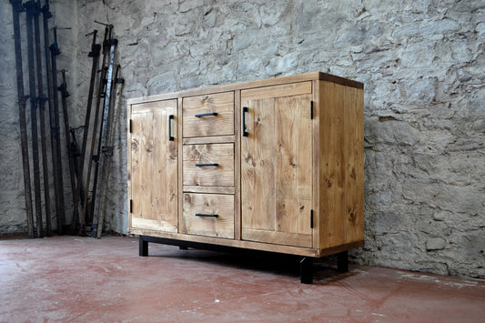 A rustic wooden sideboard cabinet with three drawers and two doors, shown in a workshop, in front of a stone wall.