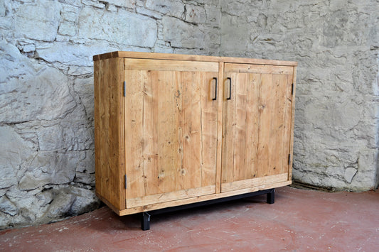 Reclaimed wood sideboard with black steel base, angled view showing two-door design and natural wood grain, photographed against a stone wall