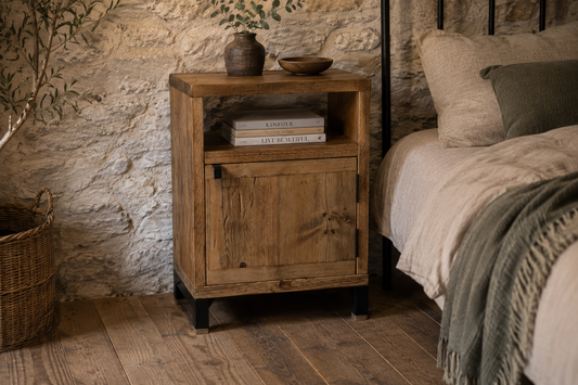A rustic wooden nightstand with a single cabinet door and open shelf, styled in a bedroom next to a bed with neutral bedding and a stone wall background.